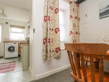 A dining area with wooden chairs and table next to a window with floral curtains and a kitchen with a washing machine and kettle at Mollys Cottage in St Just