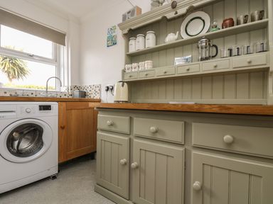 A kitchen corner with a washing machine wooden cabinets and shelves holding jars mugs and glasses at Mollys Cottage in St Just