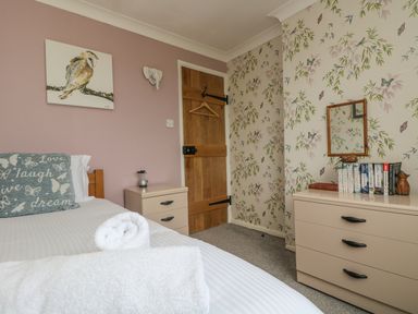 A bedroom with a bed with white linens and a pillow with text beside a wooden door a bedside table with an object and a chest of drawers with books and a mirror at Mollys Cottage in St Just