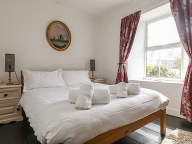 A bedroom with a double bed with white bedding and rolled towels two bedside tables with lamps a window with floral curtains and a painting on the wall at Mollys Cottage in St Just