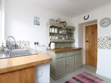A kitchen with a wooden door a gray cabinet with crockery a clock and a sink with wooden countertops at Mollys Cottage in St Just