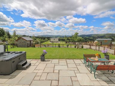 A patio with a hot tub and a table with benches overlooking a grassy yard and hills at Hillside View in Rhayader