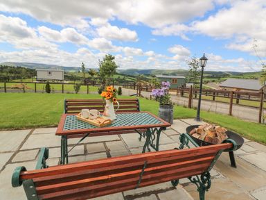 An outdoor patio with wooden benches and a metal table with flowers and cups with hills in the background at Hillside View in Rhayader