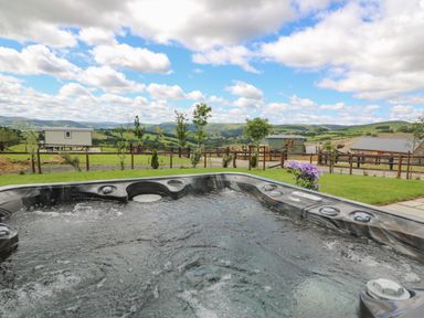 An outdoor hot tub with bubbling water overlooking a fenced grassy yard and hills in the distance at Hillside View in Rhayader