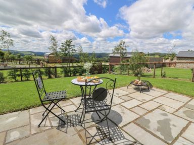 A patio with two metal chairs and a small round table with food and flowers overlooking a grassy yard and countryside at Woodland View in Rhayader