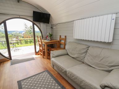 An interior room with a leather sofa wooden table with chairs flat screen tv and circular glass door showing outdoor seating at Woodland View in Rhayader