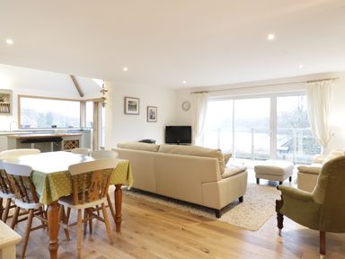 An open living room and dining area with a dining table covered with a green polka dot tablecloth chairs beige sofas an armchair a small TV and large windows with curtains at Treetops in Golant near Fowey