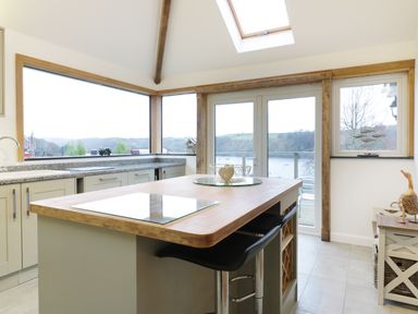 A kitchen with an island and bar stools overlooking a river through large windows at Treetops in Golant near Fowey