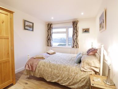 A bedroom with a double bed, wooden wardrobe, bedside table with a lamp, and window with curtains at Treetops Golant near Fowey