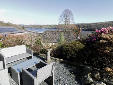 An outdoor patio with gray wicker furniture and a birdbath overlooking a lake and trees at Treetops in Golant near Fowey