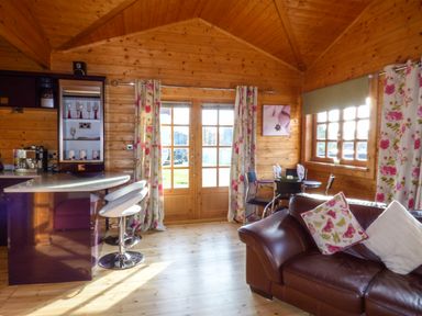 A living area with a brown leather sofa, floral curtains, a small dining table with chairs, and a kitchen counter with bar stools at St Hilda's Lodge in Hinderwell near Runswick Bay