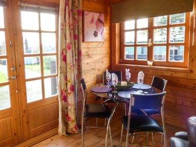 A dining area with a round glass table and black chairs near wooden windows and a door with floral curtains at St Hilda's Lodge in Hinderwell near Runswick Bay
