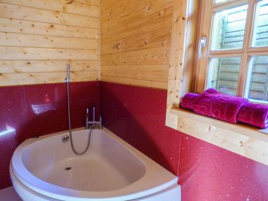 A bathroom corner with a white bathtub red tiled walls wooden paneling and a window with rolled towels on the sill at St Hildas Lodge in Hinderwell near Runswick Bay