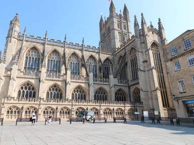 A historic stone building with large arched windows and a courtyard with benches and people in Bath