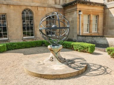 A metal armillary sphere sculpture on a stone base in a gravel courtyard with trimmed hedges and stone buildings at 33 Crescent Gardens in Bath