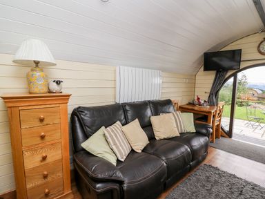 A living area with a black leather sofa with cushions a wooden chest of drawers with a lamp and sheep ornament a mounted TV and a wooden table with chairs at Mountain View in Rhayader
