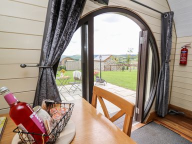 A wooden chair at a table with a bottle and snacks in a cabin with an open circular door and patio with chairs at Mountain View in Rhayader