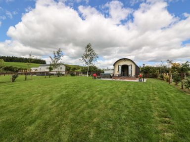 A small cabin with a patio and chairs on a grassy lawn surrounded by a fence and trees at Mountain View in Rhayader