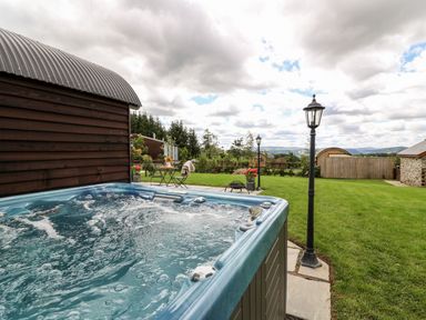 An outdoor hot tub with bubbling water next to a wooden building and a garden with two lampposts and a small table with chairs at Mountain View in Rhayader