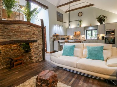 A living room with a cream sofa and turquoise cushions next to a wooden coffee table and a kitchen with grey cabinets at Elvan Cottage in Liskeard
