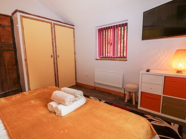 A bedroom with a bed covered by an orange blanket and towels on top a wardrobe a window with red vertical blinds a TV mounted on the wall a white storage unit with orange drawers a small round stool an orange lamp on the storage unit at Elvan Cottage in Liskeard
