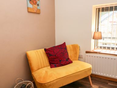 A small yellow couch with a red and black pillow next to a window with a lamp on the windowsill at Elvan Cottage in Liskeard