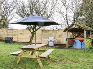 A garden with a wooden picnic table and umbrella a barbecue grill and a wooden shelter with stools at Elvan Cottage in Liskeard