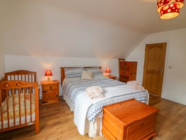 A bedroom with a double bed, a wooden crib, two bedside tables with lamps, a wooden chest, a dresser, and a wooden door at Beehive Cottage in Kilbrittain County Cork