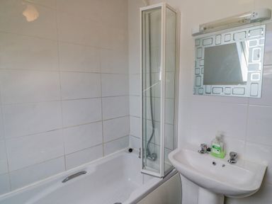 A bathroom with a bathtub a folding shower screen a sink with taps a mirror and a soap dispenser at Beehive Cottage in Kilbrittain County Cork
