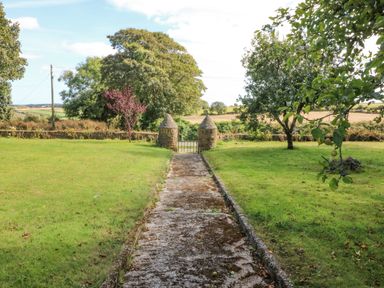 A garden with a concrete path leading to a gate between two stone pillars and trees on both sides at Beehive Cottage in Kilbrittain County Cork