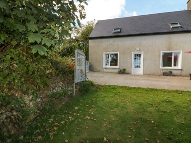 A yard with grass and a paved patio outside a house with two windows and a white door at Beehive Cottage in Kilbrittain County Cork