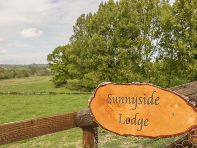 A wooden sign reading Sunnyside Lodge on a fence with green fields and trees in the background at Sunnyside Lodge in Crewkerne