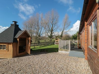 An outdoor space with a wooden cabin with chimney a gravel area a wooden deck and trees in the background at Sunnyside Lodge in Crewkerne