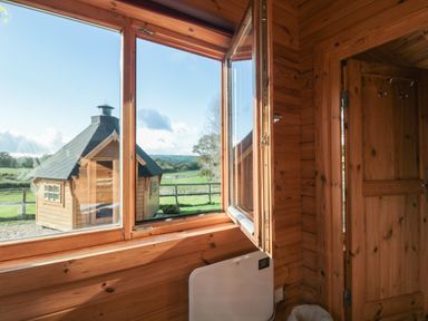 A wooden interior with an open window showing a wooden cabin and green fields outside at Sunnyside Lodge Crewkerne