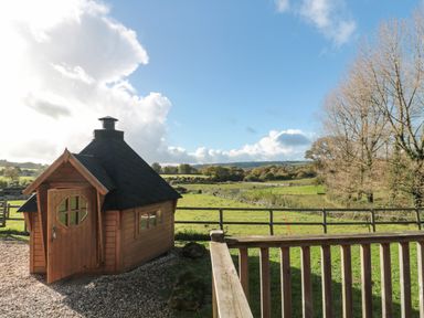 A wooden cabin on gravel next to a fenced green field with trees and hills at Sunnyside lodge in Crewkerne