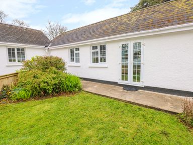A white exterior wall with windows and a glass door next to a garden with grass and bushes at 4 Hanbury Lodge in Pentlepoir
