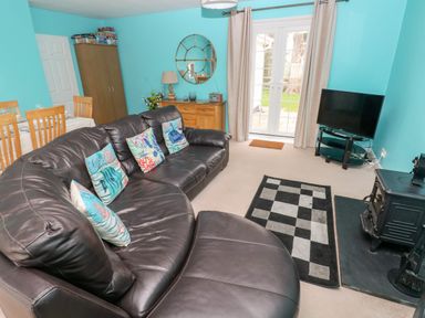 A living room with a brown leather sectional sofa and patterned cushions a wooden dining table with chairs a wooden cabinet a round mirror and a television on a stand at 4 Hanbury Lodge Pentlepoir