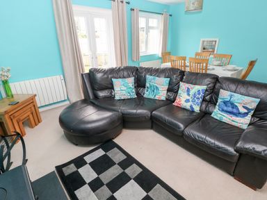 A living room with a black leather sectional sofa with sea-themed pillows a dining table with wooden chairs and tableware blue walls and beige curtains at 4 Hanbury Lodge in Pentlepoir