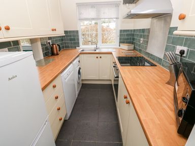 A kitchen with wooden countertops, white cabinets, a refrigerator, washing machine, dishwasher, sink under a window, stove, microwave, kettle, and knife block at 4 Hanbury Lodge in Pentlepoir