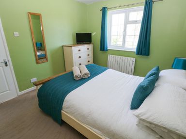 A bedroom with a bed with teal and white bedding a dresser with a TV a window with teal curtains and a wall mirror at 4 Hanbury Lodge in Pentlepoir