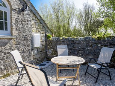 An outdoor patio with a round wooden table and four mesh chairs by a stone wall and house at Moor View Chapel in Camelford