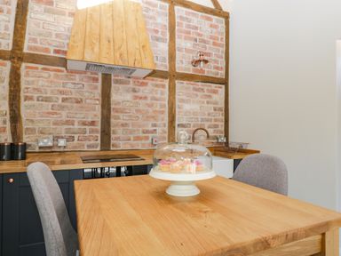 A kitchen with a wooden table two gray chairs a cake stand with cupcakes and a brick wall at The Stables in Cross Keys near Hereford