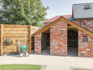 An outdoor bike and bin store made of brick with a wooden fence holding a dog waste bin and garden hose at The Stables in Cross Keys near Hereford