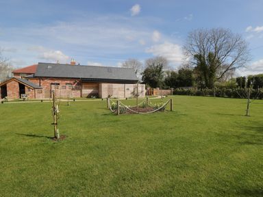 A large green lawn with young trees and a building with brick and wooden walls in the background at The Stables in Cross Keys near Hereford