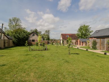 A grassy garden area with small trees and a pathway surrounded by houses at The Stables in Cross Keys near Hereford