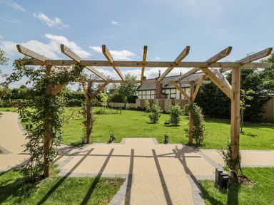 A wooden pergola on a paved path in a garden with grass and shrubs at The Stables in Cross Keys near Hereford