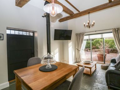 A dining area with a wooden table, chairs, a wall-mounted television, and a sitting area with a coffee table and sofa with sliding glass doors to a patio at The Stables in Cross Keys near Hereford