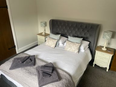 A bedroom with a bed with white bedding gray headboard two bedside tables with lamps and folded towels at The Stables in Cross Keys near Hereford