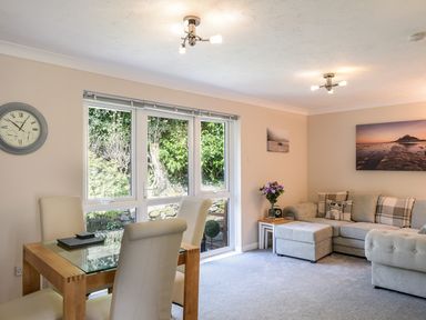 A living room with a glass dining table and white chairs next to a beige sofa with cushions and a side table with flowers at Little Retreat in Liskeard