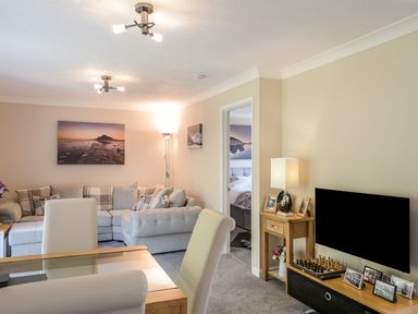 A living room with a grey sectional sofa a glass dining table with chairs a wooden TV stand with a television and framed photos at Little Retreat in Liskeard
